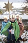 Quelle: © Mika Väisänen / M@RS MediaPortal Sternsinger Vier Kinder mit goldenen Kronen und bunten Umhängen stehen im Schnee, eines hält einen großen goldenen Stern auf einer Stange und eine Spendendose